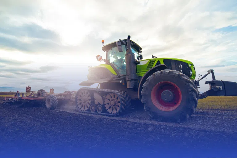 A large green tractor with tracks, instead of wheels, is plowing a field under a partly cloudy sky. The sun is visible in the background, casting a bright light over the scene.
