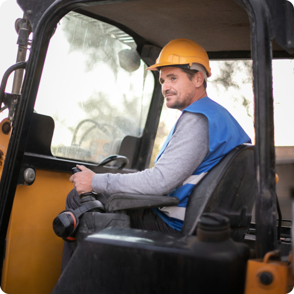 Male crane operator wearing a yellow hard hat and blue safety vest inside the control cabin of construction equipment