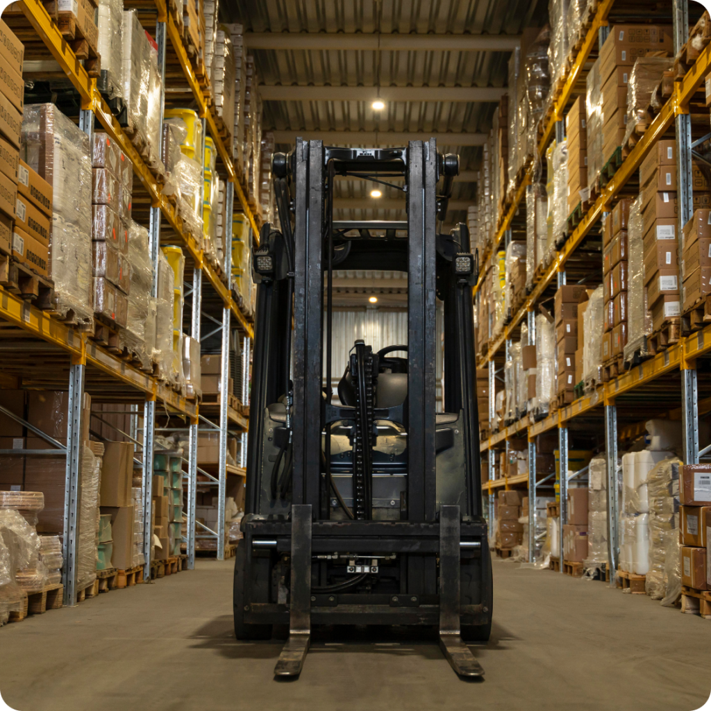 Front view of a forklift positioned between tall warehouse shelves filled with stacked boxes and pallets, ready for inventory handling and storage operations