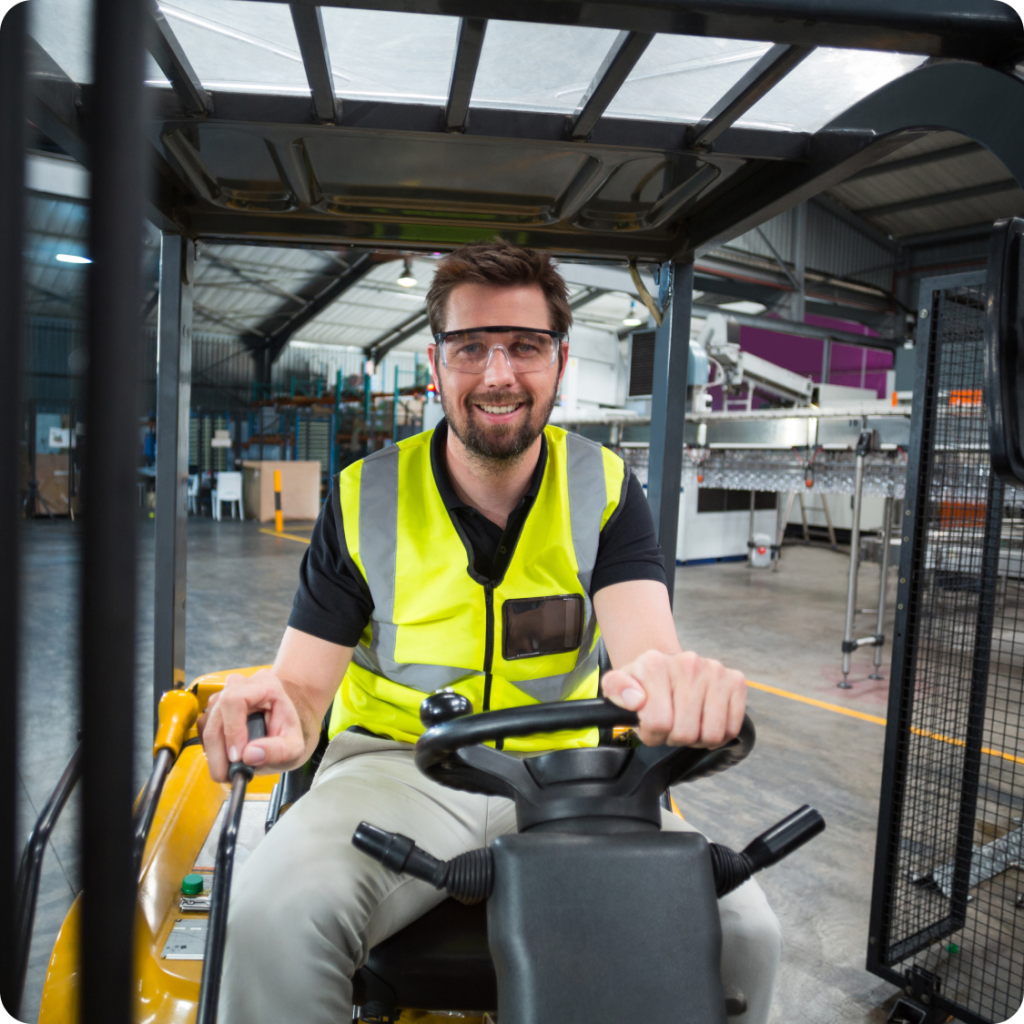 Smiling forklift operator wearing a high-visibility vest and safety glasses, operating a lift truck inside a modern warehouse