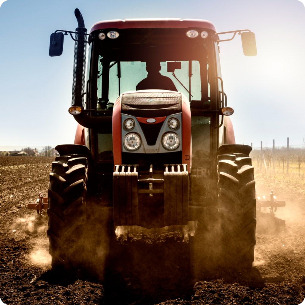 Front view of a modern red tractor plowing dry soil in a field with sunlight in the background and a farmer silhouette visible inside the cabin