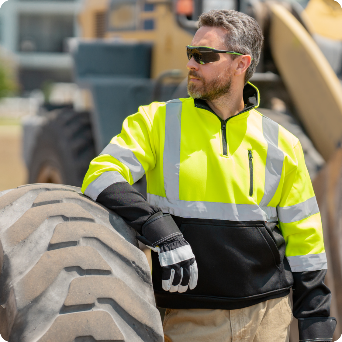 Construction worker in yellow high-visibility jacket and gloves leaning on large heavy machinery tire outdoors