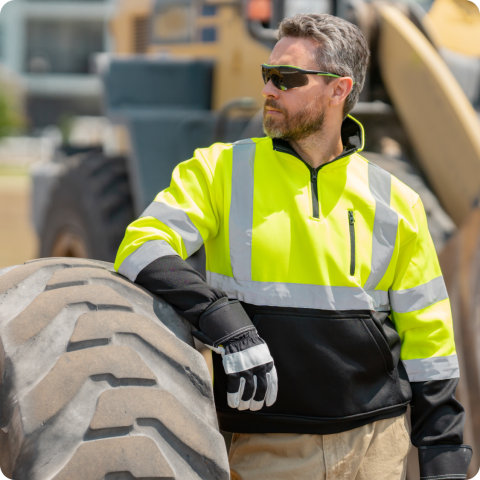 Construction Worker Wearing High-Visibility Safety Gear Near Heavy Machinery Tire Construction worker in yellow high-visibility jacket and gloves leaning on large heavy machinery tire outdoors