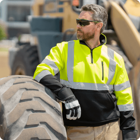 Construction worker in yellow high-visibility jacket and gloves leaning on large heavy machinery tire outdoors